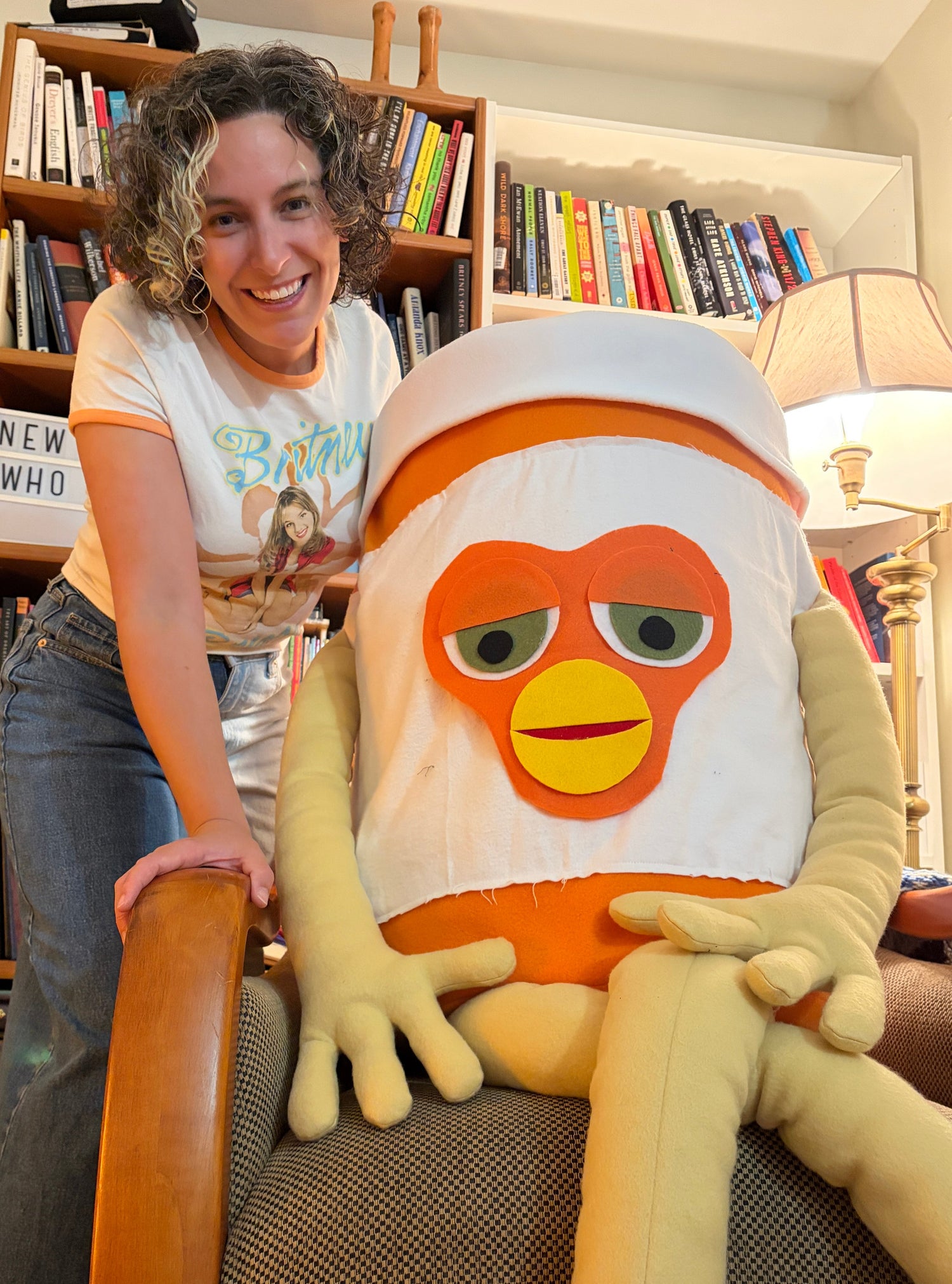 Emma, a white woman with curly brown hair, stands next to a large pill bottle stuffed creature with a furby face.
